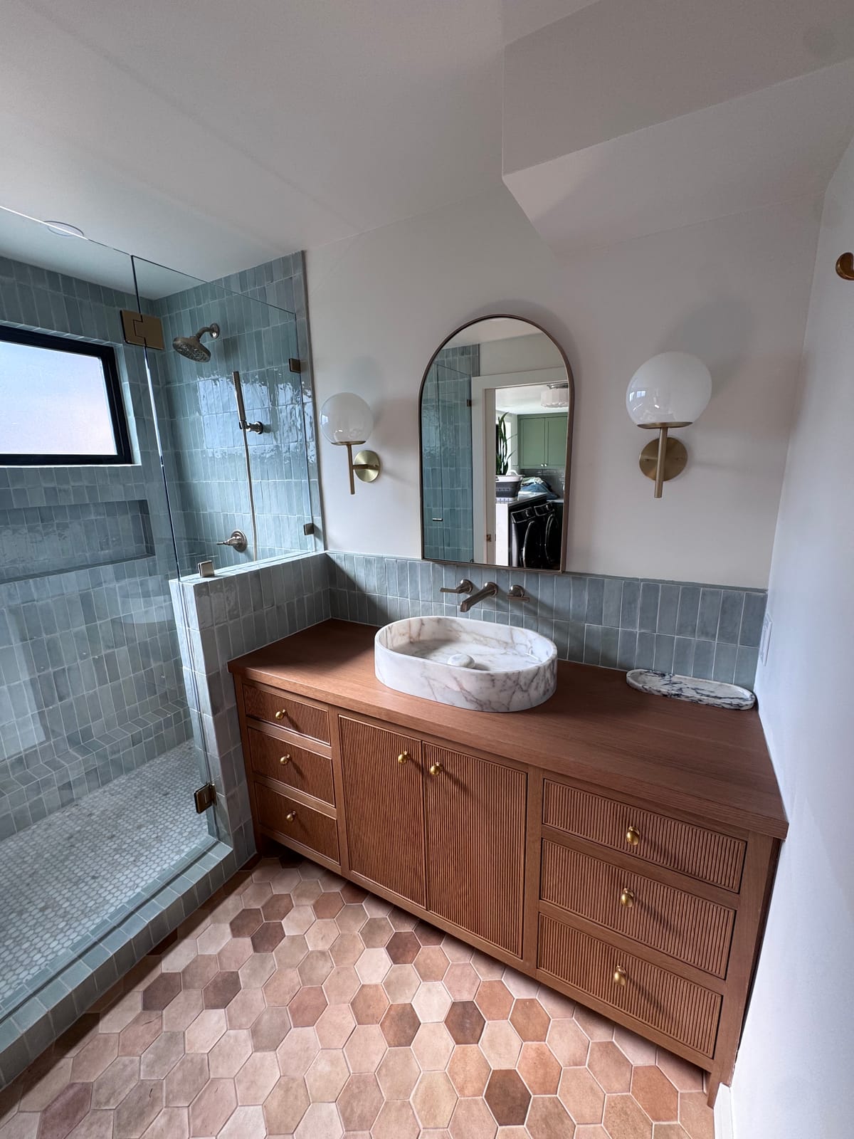 Finished bathroom remodel in Whitford near Tigard — wood slat vanity, marble vessel sink, arched mirror, and brass sconces beside the glazed green tile shower — K&H Construction
