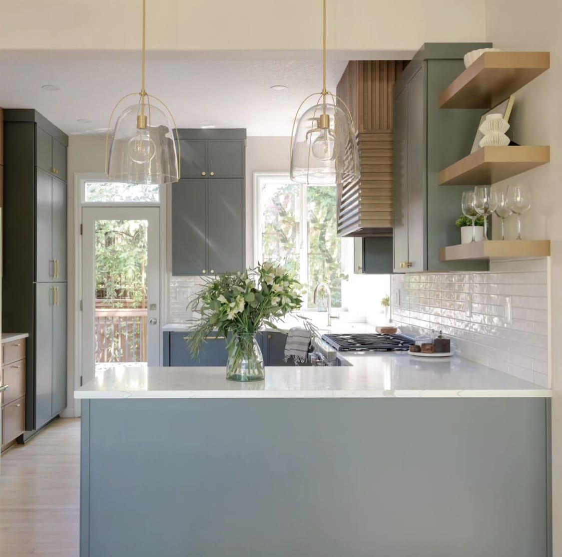 After the remodel: sage-green cabinetry, white quartz peninsula, brass-and-glass pendants, and a custom fluted wood range hood in the same Ashcreek kitchen — K&H Construction